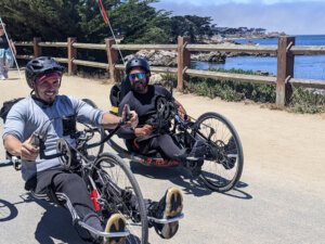 two young men on hand cycles are smiling while riding by a fence. the ocean is behind them