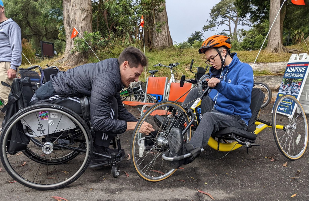 one person in a wheelchair is adjusting the foot pedals for another person who in an a hand tricycle