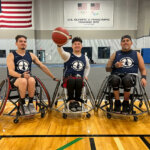 Three men in wheelchair basketball chairs wearing National Wheelchair Basketball Association shirts (left to right, Jorge, Artur, and Armando) pose on the court with a basketball.