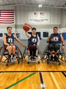 Three men in wheelchair basketball chairs wearing National Wheelchair Basketball Association shirts (left to right, Jorge, Artur, and Armando) pose on the court with a basketball.