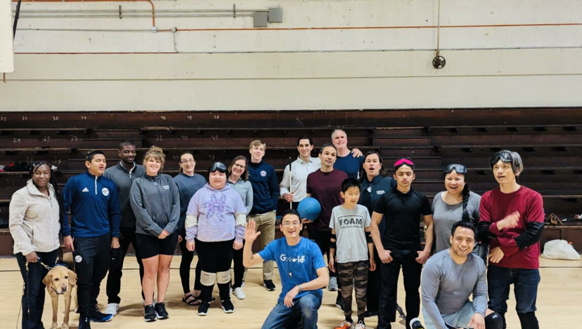 A large, diverse group of people, including members of BORP's goalball team and the USA Paralympic Goalball Team, pose together in an indoor gymnasium. They are smiling and appear to be in good spirits, celebrating a successful and engaging practice session. Some are wearing goalball gear like sleep shades or kneepads, and a guide dog is also present in the photo.