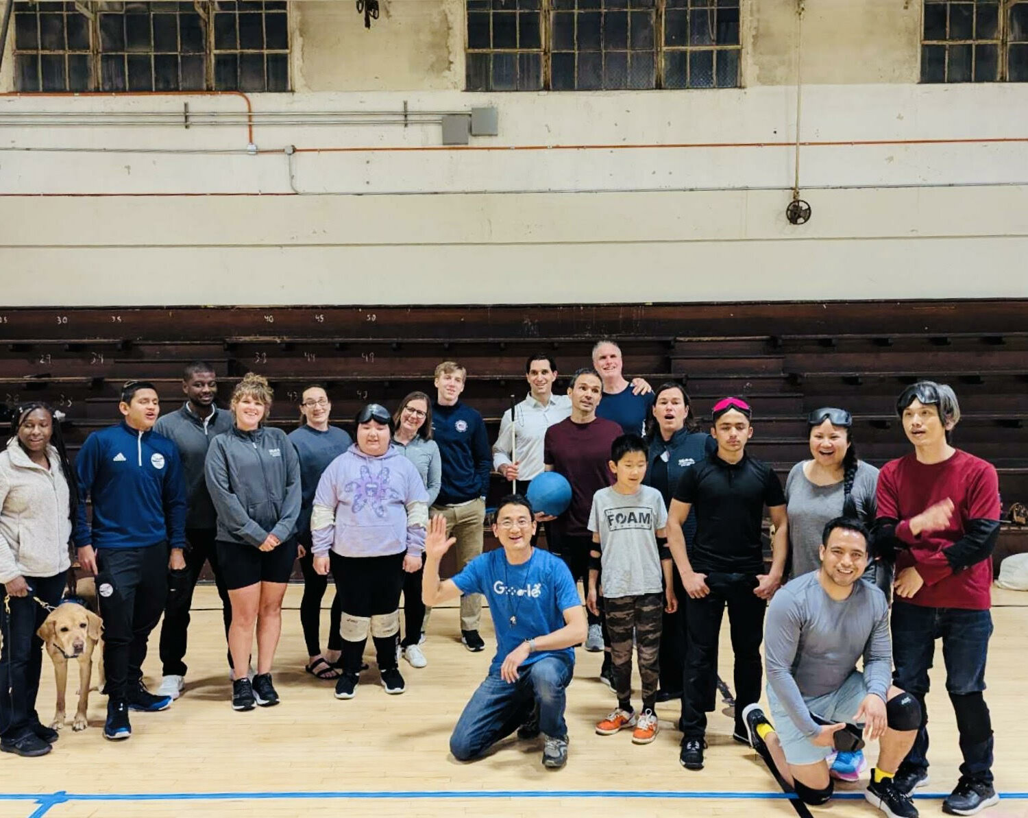 A large, diverse group of people, including members of BORP's goalball team and the USA Paralympic Goalball Team, pose together in an indoor gymnasium. They are smiling and appear to be in good spirits, celebrating a successful and engaging practice session. Some are wearing goalball gear like sleep shades or kneepads, and a guide dog is also present in the photo.