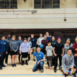 A large, diverse group of people, including members of BORP's goalball team and the USA Paralympic Goalball Team, pose together in an indoor gymnasium. They are smiling and appear to be in good spirits, celebrating a successful and engaging practice session. Some are wearing goalball gear like sleep shades or kneepads, and a guide dog is also present in the photo.