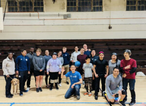 A large, diverse group of people, including members of BORP's goalball team and the USA Paralympic Goalball Team, pose together in an indoor gymnasium. They are smiling and appear to be in good spirits, celebrating a successful and engaging practice session. Some are wearing goalball gear like sleep shades or kneepads, and a guide dog is also present in the photo.