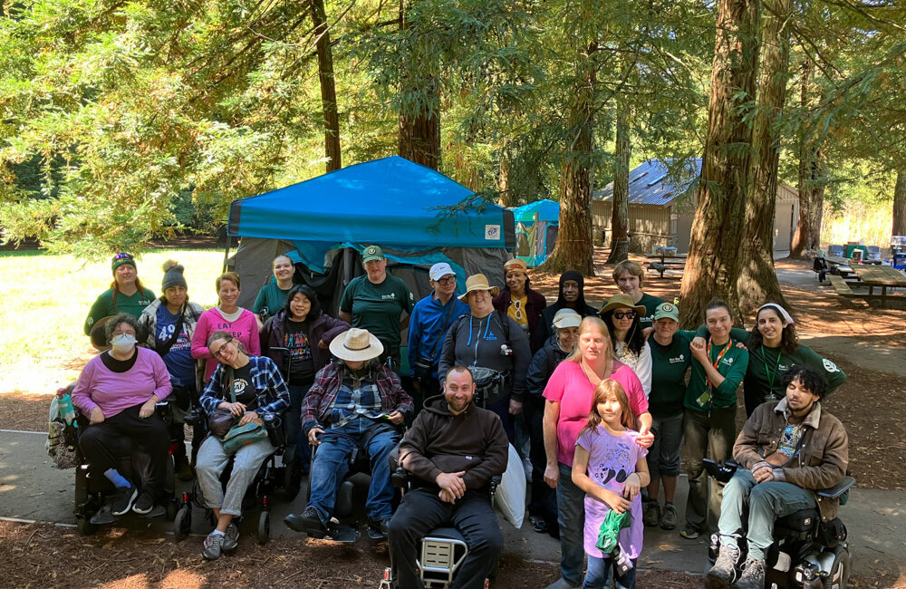 Image1: 23 people gathered at a campsite, five are in wheelchairs and at least 6 are wearing green East Bay Parks t-shirts.