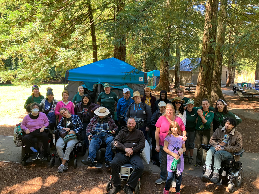 Image1: 23 people gathered at a campsite, five are in wheelchairs and at least 6 are wearing green East Bay Parks t-shirts.