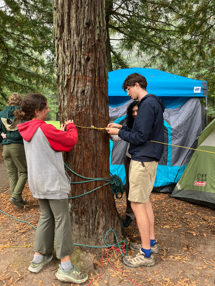 Image 2: 3 volunteers help secure the guide ropes for blind campers to be able to find their way to the bathroom at night. The guide ropes went from the tents of each blind camper to the bathroom, with knots to indicate which tent was which so they got back to the right sleeping bag. Participants learned other tips for accessible camping such as how to set up a tent with enough internal space and ways to get the bed high enough off the ground for wheelchair users.