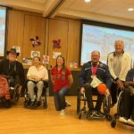 A group of 6 paralympians and olympians are lined up smiling at the camera, four in their wheelchairs and two kneeling.