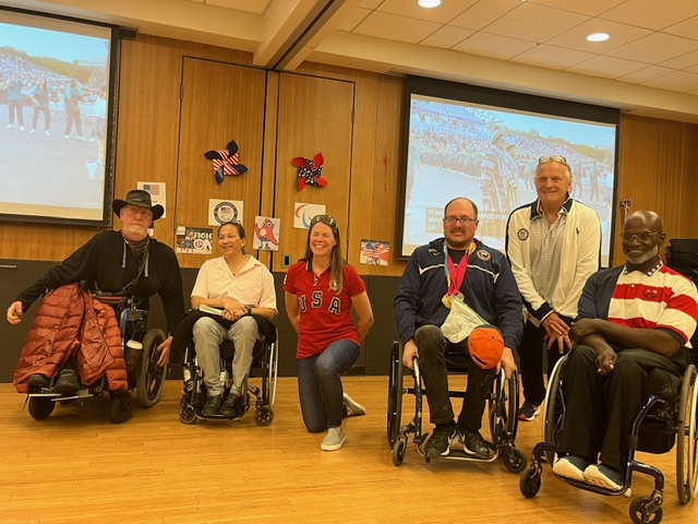 A group of 6 paralympians and olympians are lined up smiling at the camera, four in their wheelchairs and two kneeling.