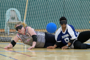 Two female goalball players, one in a gray jersey and one in a blue jersey, lie on the floor mid-play with a blue ball between them. Both are wearing blackout goggles, and a net is visible behind them.