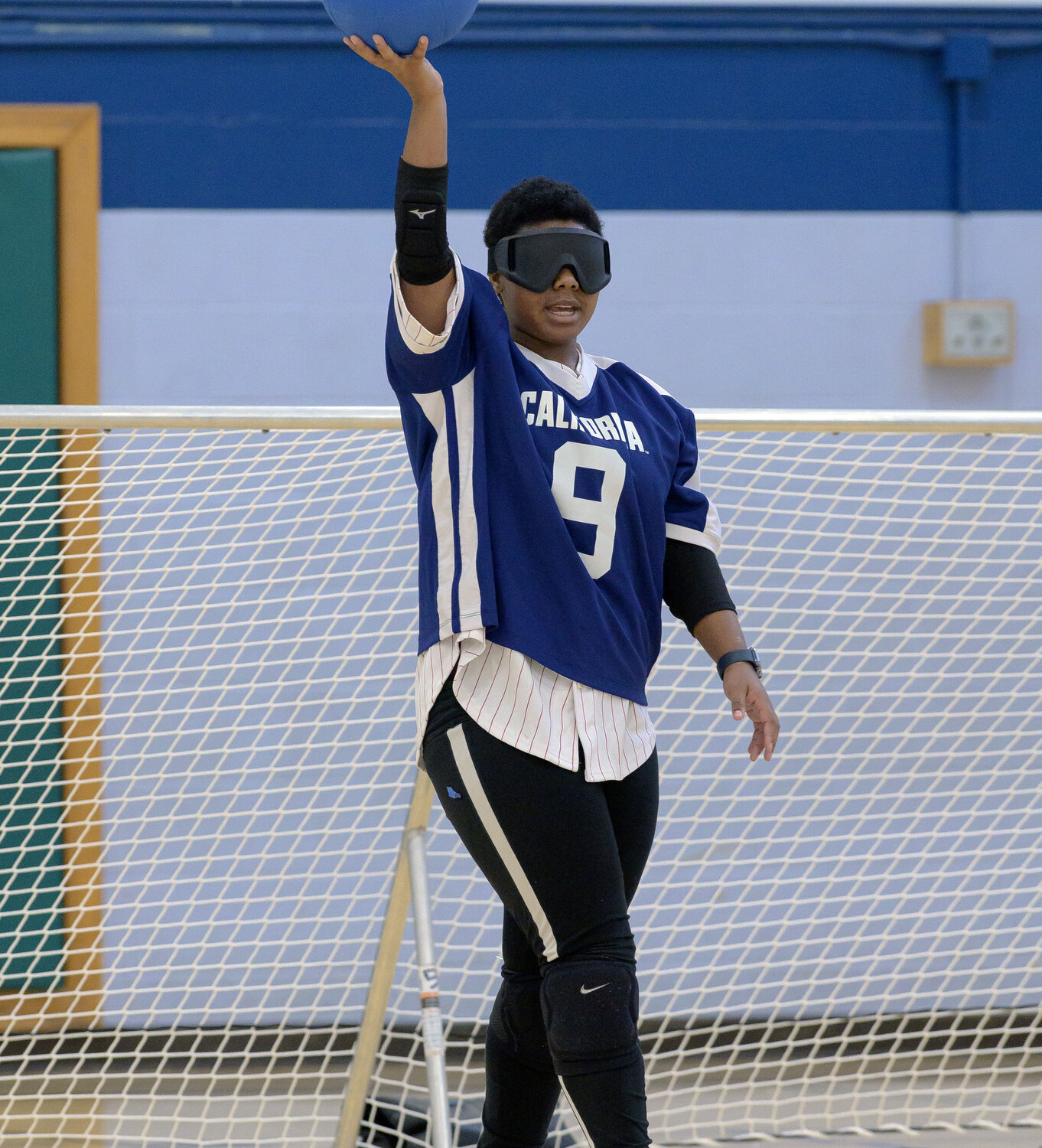 A goalball player in a blue jersey with the number 9 holds a blue ball aloft with one hand, preparing to throw. The player wears blackout goggles and stands in front of a goal net.