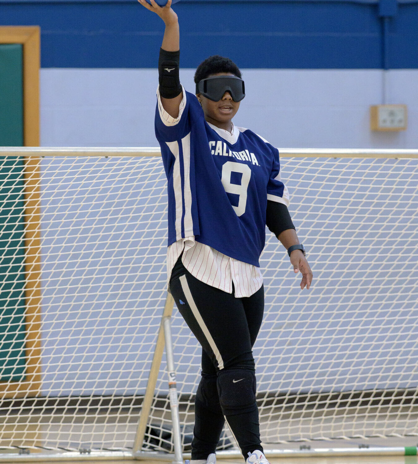 A goalball player in a blue jersey with the number 9 holds a blue ball aloft with one hand, preparing to throw. The player wears blackout goggles and stands in front of a goal net.