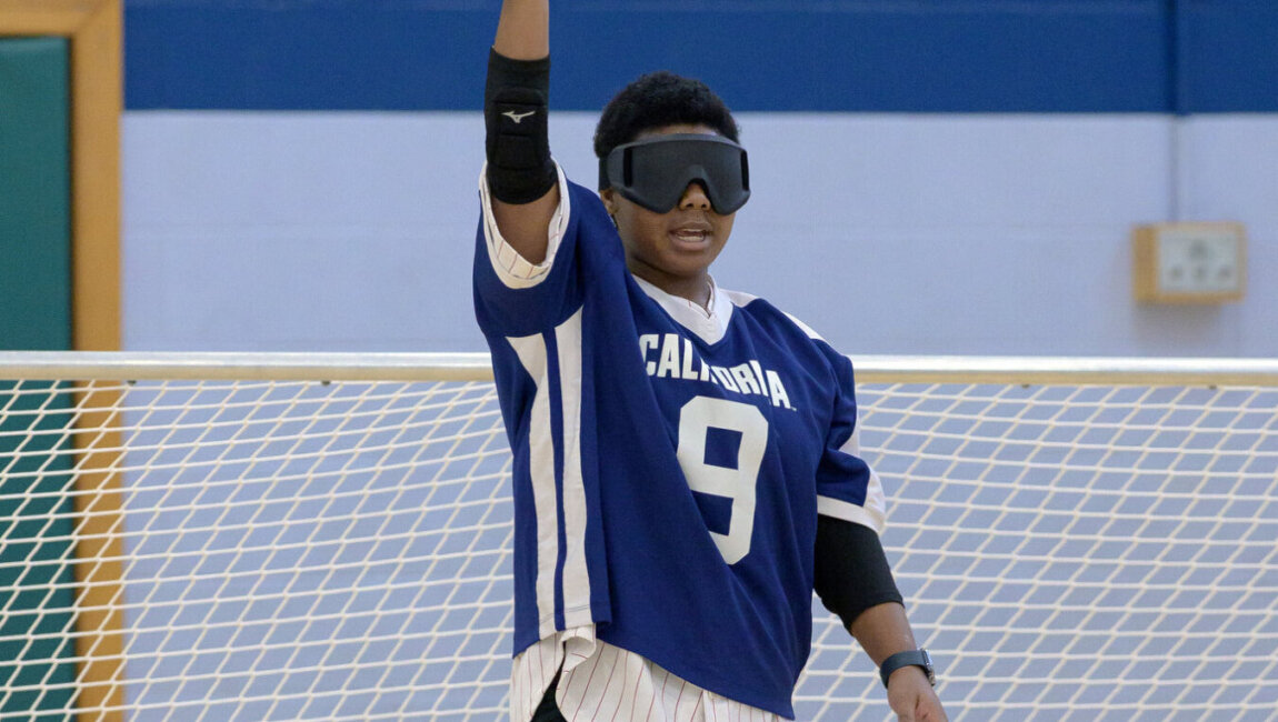 A goalball player in a blue jersey with the number 9 holds a blue ball aloft with one hand, preparing to throw. The player wears blackout goggles and stands in front of a goal net.