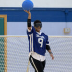 A goalball player in a blue jersey with the number 9 holds a blue ball aloft with one hand, preparing to throw. The player wears blackout goggles and stands in front of a goal net.