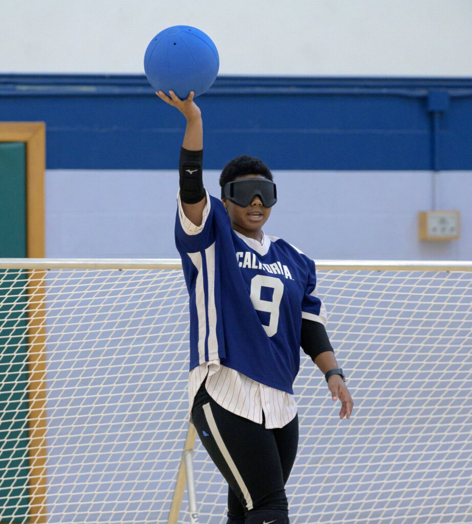 A goalball player in a blue jersey with the number 9 holds a blue ball aloft with one hand, preparing to throw. The player wears blackout goggles and stands in front of a goal net.