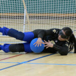 A goalball player with long hair in a ponytail in a black jersey with gold accents lies on the court holding a blue ball. The player wears blackout goggles, with a goal net visible behind them.