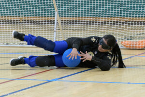 A goalball player with long hair in a ponytail in a black jersey with gold accents lies on the court holding a blue ball. The player wears blackout goggles, with a goal net visible behind them.