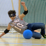 A goalball player in a gray jersey with "California" and the number 10 printed on it is mid-slide, attempting to block the blue ball. The player wears blackout goggles and is on an indoor court.