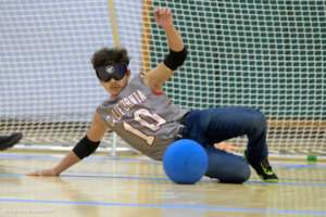 A goalball player in a gray jersey with "California" and the number 10 printed on it is mid-slide, attempting to block the blue ball. The player wears blackout goggles and is on an indoor court.