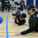 Two goalball players in black jerseys dive on the court, reaching to block a blue ball. Other players and spectators are visible in the background.