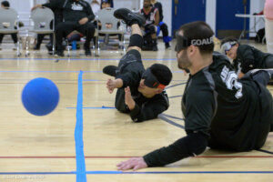 Two goalball players in black jerseys dive on the court, reaching to block a blue ball. Other players and spectators are visible in the background.