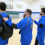 Three players wearing bright blue jerseys stand in a line during goalball practice, with their hands on each other's shoulders. The first player carries a black backpack with a water bottle. A goal net is visible in the background, and the setting is an indoor gymnasium.