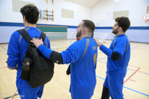 Three players wearing bright blue jerseys stand in a line during goalball practice, with their hands on each other's shoulders. The first player carries a black backpack with a water bottle. A goal net is visible in the background, and the setting is an indoor gymnasium.