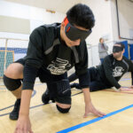 Two players wearing black jerseys crouch on the court during goalball practice. They wear blackout goggles, and a goal net is visible behind them. One player is utilizing the tactile markings on the floor for orientation