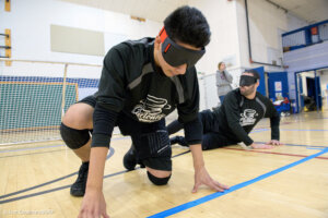 Two players wearing black jerseys crouch on the court during goalball practice. They wear blackout goggles, and a goal net is visible behind them. One player is utilizing the tactile markings on the floor for orientation