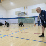 A goalball referee observes players crawling on the court during a training session. The players, dressed in black jerseys, wear blackout goggles and are positioned near a goal net. A woman in casual clothing stands in the background, watching the activity.