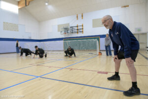 A goalball referee observes players crawling on the court during a training session. The players, dressed in black jerseys, wear blackout goggles and are positioned near a goal net. A woman in casual clothing stands in the background, watching the activity.