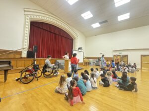 Students sit on a gym floor watching a presentation on adaptive sports, with a wheelchair and speakers visible in the foreground.