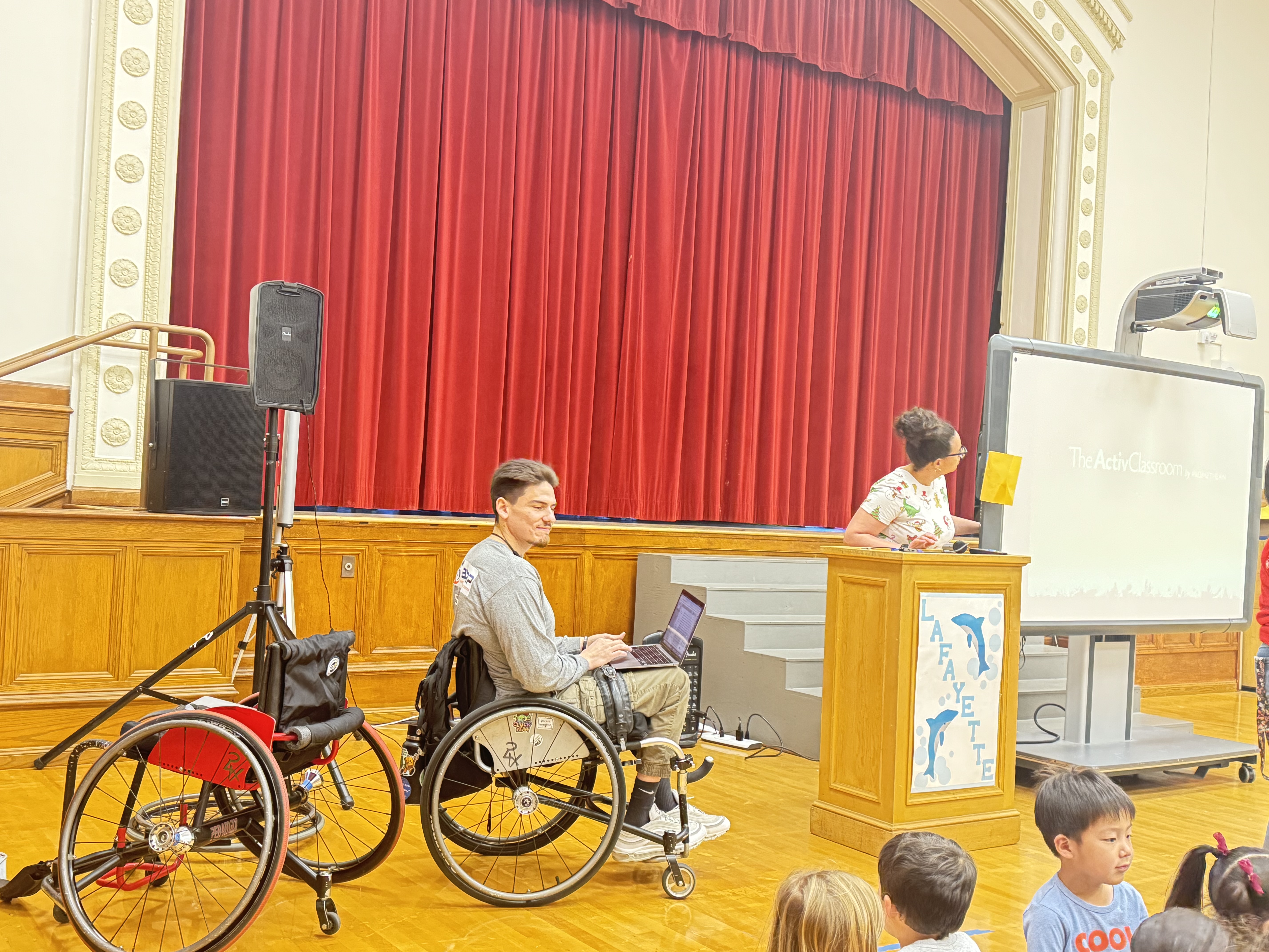 A man in wheelchair sits at the front of an assembly room and addresses a group of young students, several of whom can be seen in the background. A sports chair is next to him.