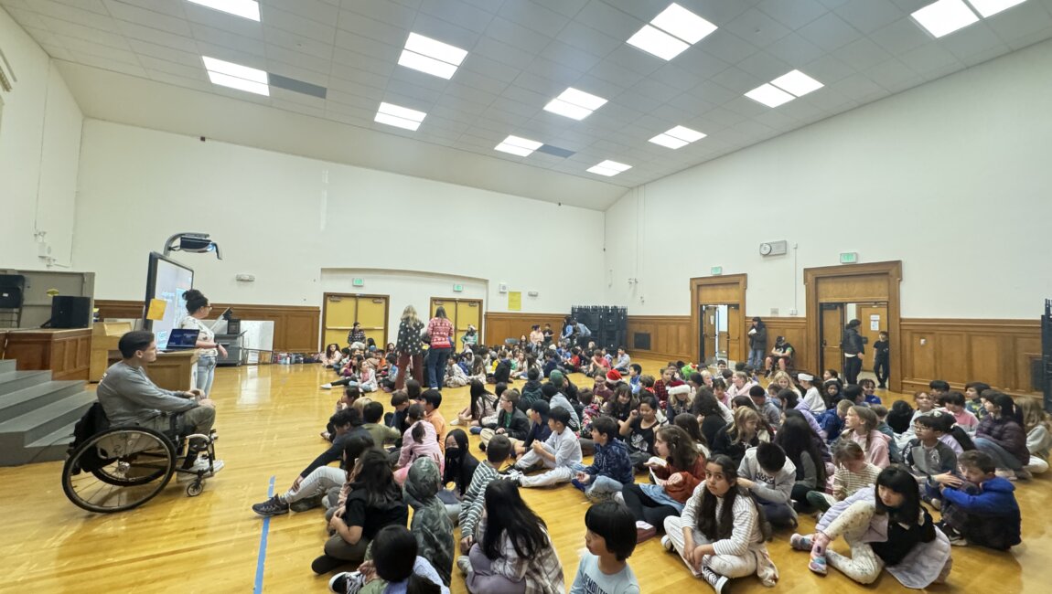 a large group of elementary school students sit in a gy, listening to a presentation led by a man in a wheelchair at the front of the room.