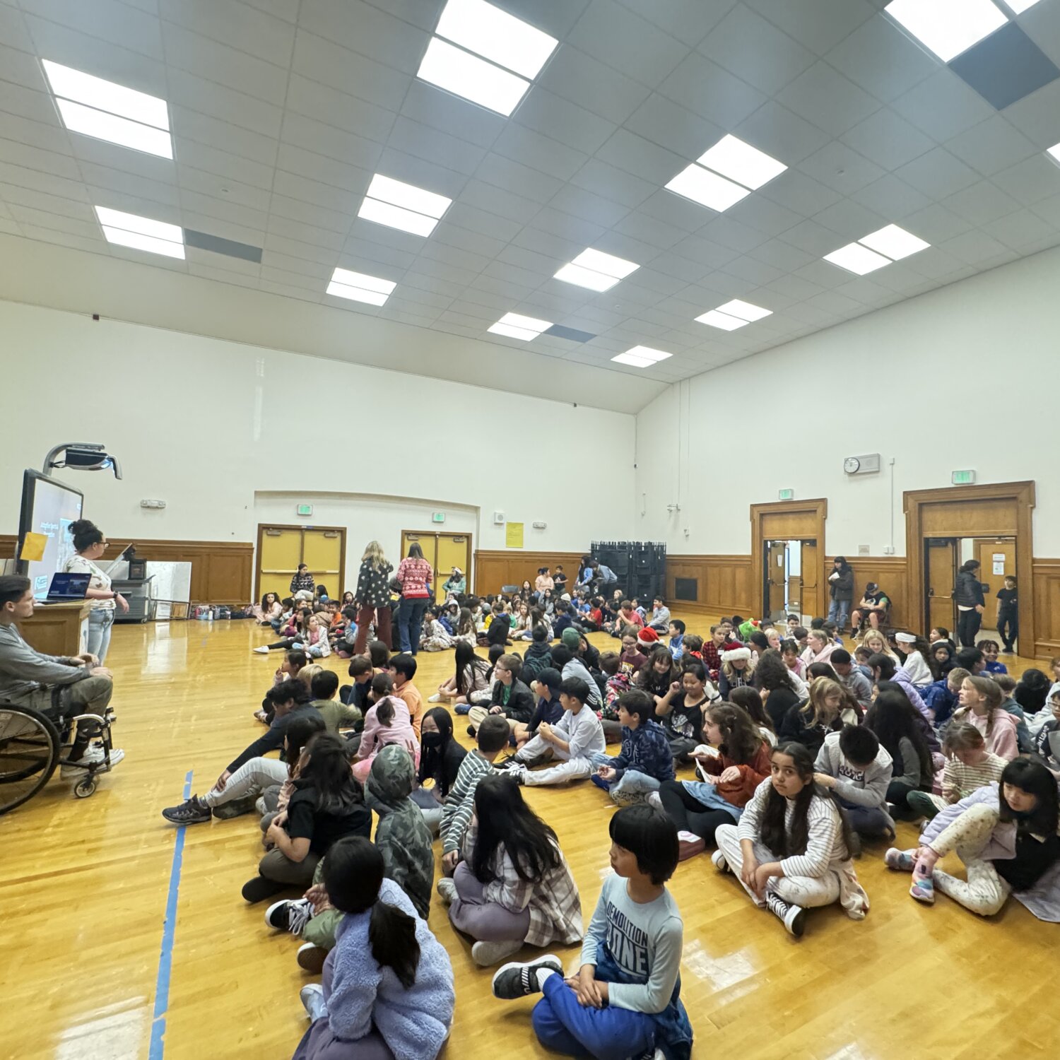 a large group of elementary school students sit in a gy, listening to a presentation led by a man in a wheelchair at the front of the room.