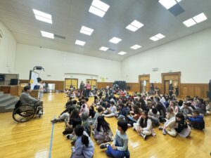 a large group of elementary school students sit in a gy, listening to a presentation led by a man in a wheelchair at the front of the room.