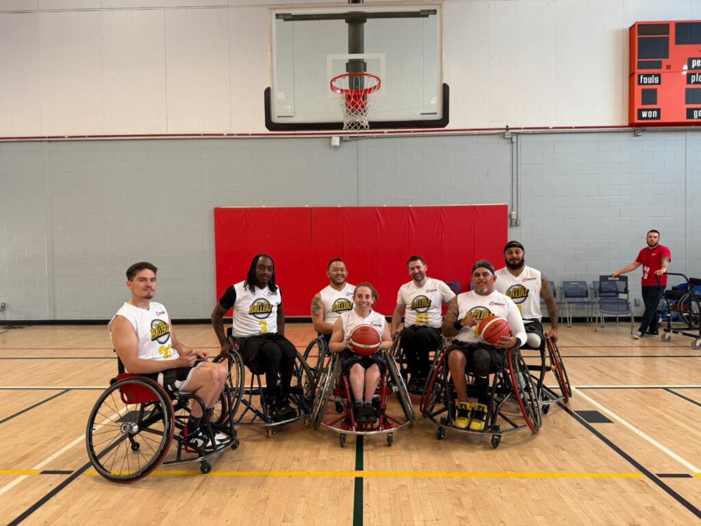 A group of seven wheelchair basketball players posing for a photo on an indoor basketball court. They are seated in their sports wheelchairs, holding basketballs and wearing matching white jerseys with the team logo and numbers. Behind them, a basketball hoop and red padding are visible on the wall. Two individuals in casual clothing stand near the court in the background, one giving a thumbs-up. The setting is a brightly lit gymnasium with wooden flooring and a scoreboard on the wall.