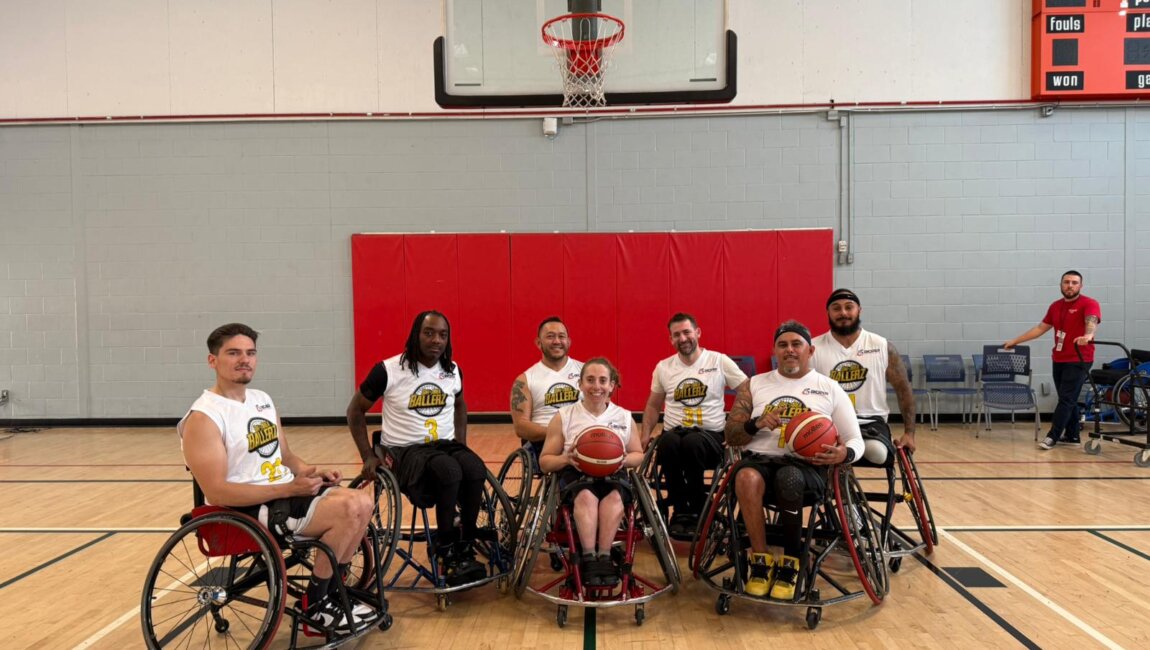 A group of seven wheelchair basketball players posing for a photo on an indoor basketball court. They are seated in their sports wheelchairs, holding basketballs and wearing matching white jerseys with the team logo and numbers. Behind them, a basketball hoop and red padding are visible on the wall. Two individuals in casual clothing stand near the court in the background, one giving a thumbs-up. The setting is a brightly lit gymnasium with wooden flooring and a scoreboard on the wall.