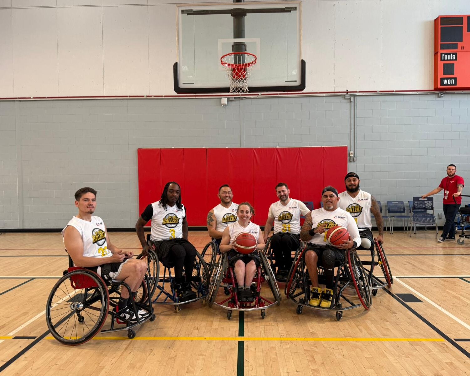 A group of seven wheelchair basketball players posing for a photo on an indoor basketball court. They are seated in their sports wheelchairs, holding basketballs and wearing matching white jerseys with the team logo and numbers. Behind them, a basketball hoop and red padding are visible on the wall. Two individuals in casual clothing stand near the court in the background, one giving a thumbs-up. The setting is a brightly lit gymnasium with wooden flooring and a scoreboard on the wall.