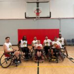 A group of seven wheelchair basketball players posing for a photo on an indoor basketball court. They are seated in their sports wheelchairs, holding basketballs and wearing matching white jerseys with the team logo and numbers. Behind them, a basketball hoop and red padding are visible on the wall. Two individuals in casual clothing stand near the court in the background, one giving a thumbs-up. The setting is a brightly lit gymnasium with wooden flooring and a scoreboard on the wall.