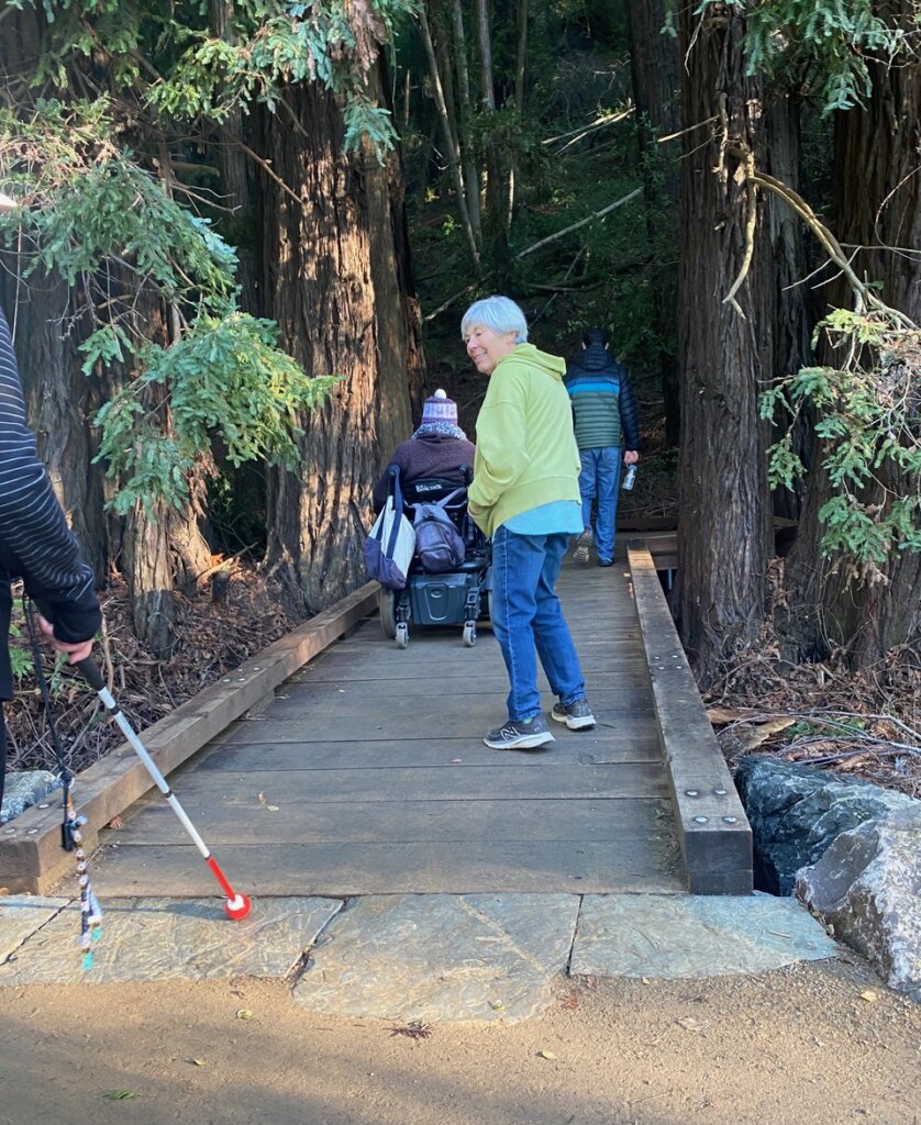 Four people, one in a power chair, are crossing a flat accessible wooden boardwalk deeper into the redwood forest.