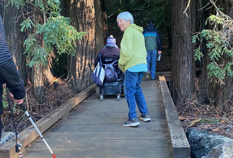 Four people, one in a power chair, are crossing a flat accessible wooden boardwalk deeper into the redwood forest.