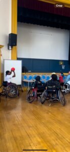 A group of 6 elementary school students sit in wheelchairs. One holds a ball and prepares to pass. A small portable net is seen in background