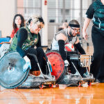 A fierce wheelchair rugby matchup unfolds as two competitors in gray and green jerseys push their specialized sports chairs forward. A referee in black observes closely, while another player watches in the background.