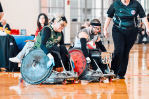 A fierce wheelchair rugby matchup unfolds as two competitors in gray and green jerseys push their specialized sports chairs forward. A referee in black observes closely, while another player watches in the background.