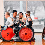 A group of wheelchair rugby players engages in a match on a polished indoor court. Two players in gray jerseys push aggressively toward an opponent in a green and gold jersey. Their red-wheeled sports chairs are reinforced with protective guards.