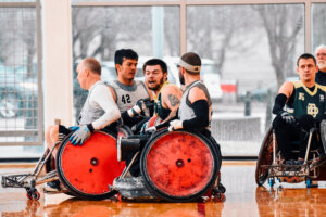 A group of wheelchair rugby players engages in a match on a polished indoor court. Two players in gray jerseys push aggressively toward an opponent in a green and gold jersey. Their red-wheeled sports chairs are reinforced with protective guards.