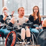 A red-haired athlete in a white and black wheelchair rugby jersey (#9) sits in a sports wheelchair, gripping the wheels with gloved hands. Behind him, a woman claps while another spectator watches attentively. The indoor court has large windows showing raindrops on the glass.