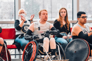 A red-haired athlete in a white and black wheelchair rugby jersey (#9) sits in a sports wheelchair, gripping the wheels with gloved hands. Behind him, a woman claps while another spectator watches attentively. The indoor court has large windows showing raindrops on the glass.