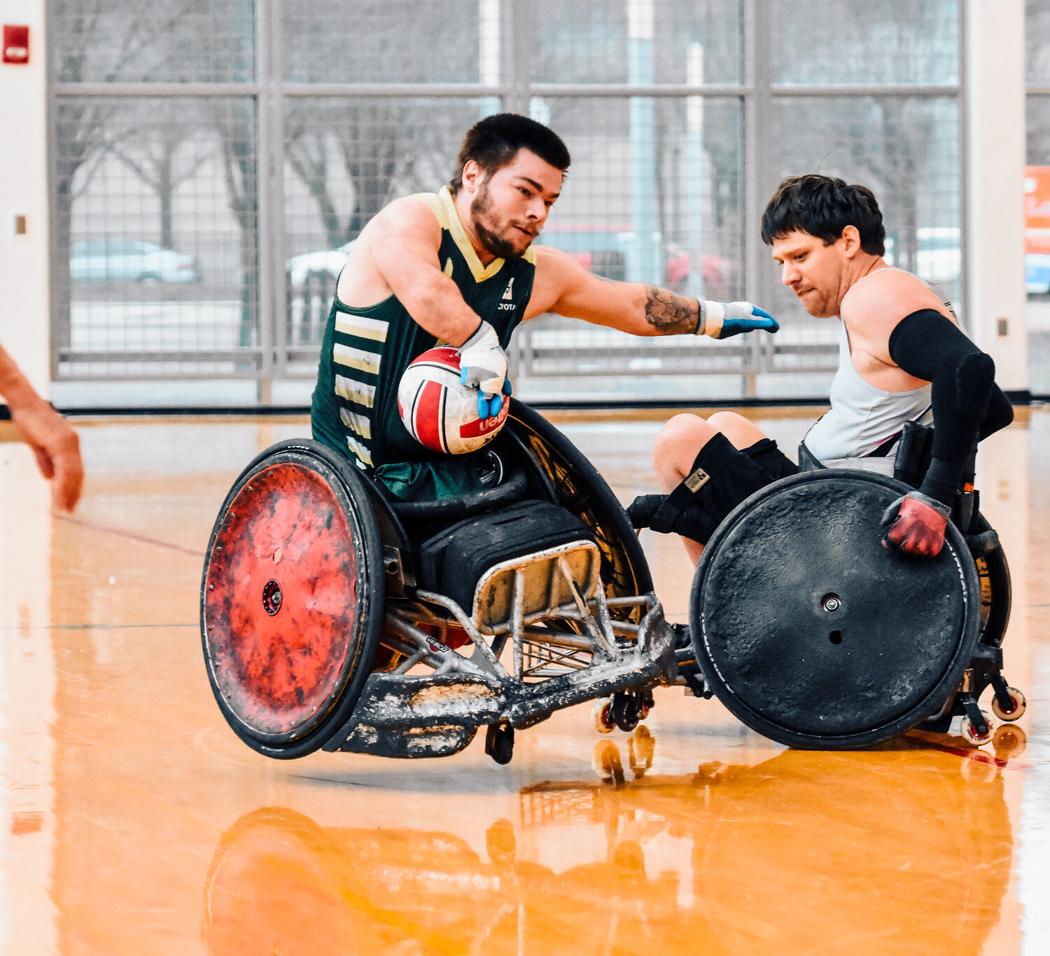 A player in a green and gold jersey grips the ball tightly while an opponent in a gray jersey attempts to block him. Both athletes are in motion, their sports wheelchairs colliding on the polished court.